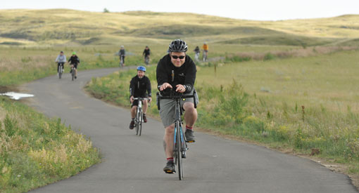 Bikers cycle along the Badlands Parkway, scenic grassy hills in the background.