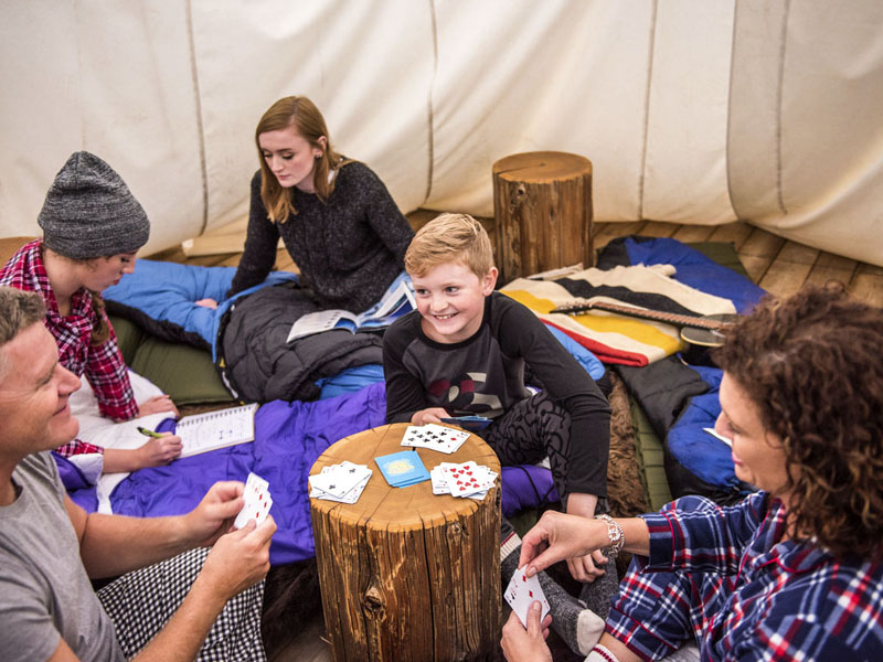A family dressed in pajamas plays a game of cards while camping overnight inside a tipi.