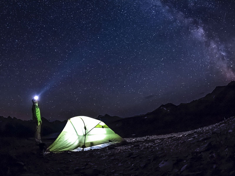 A man with a headlamp stands by his tent with the Milky Way visible in the background.