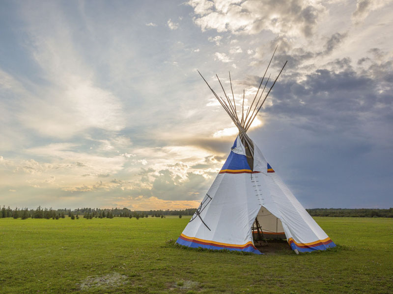 A tipi on large field under a cloudy sky with some rays of sun peaking through.