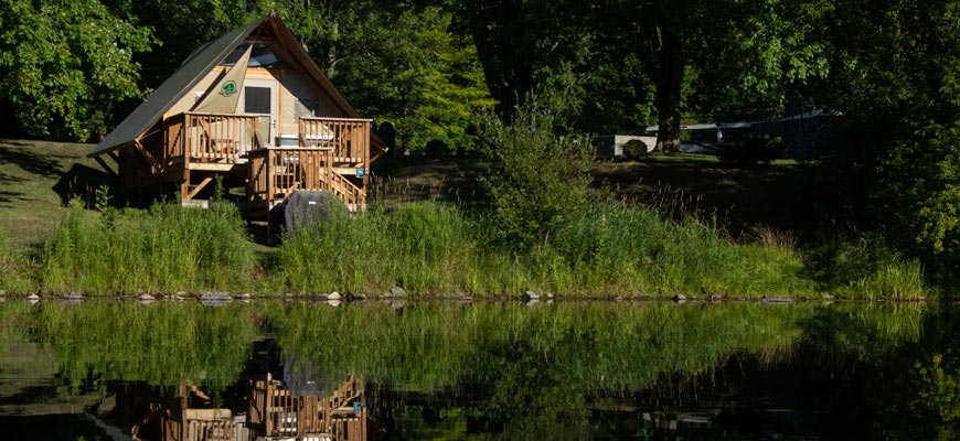 An oTENTik tent stands on the shore high above a clump of cattails with its image reflected in water