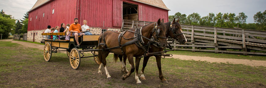 A team of horses pulls a wagon full of visitors past a big red barn.