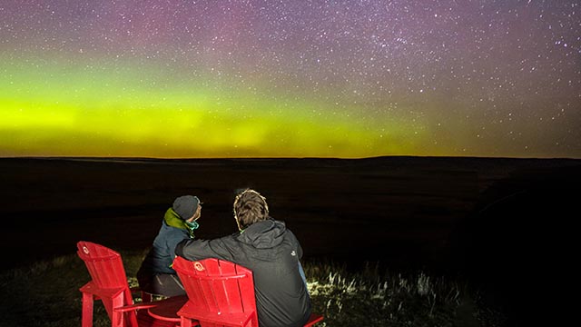 Two visitors enjoying the breathtaking views of the Milky Way and bright green and purple Aurora Borealis from the comforts of red chairs at night in Grasslands National Park.