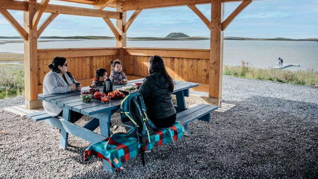 Two women and two young children sit at a picnic table in a shelter by the ocean with a pingo in the background at the Pingo Canadian Landmark.