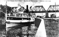 The S.S. Empress with many passengers on the bridge leaving the Sainte-Anne-de-Bellevue lock towards the Ottawa River. In the background, a railway bridge. 