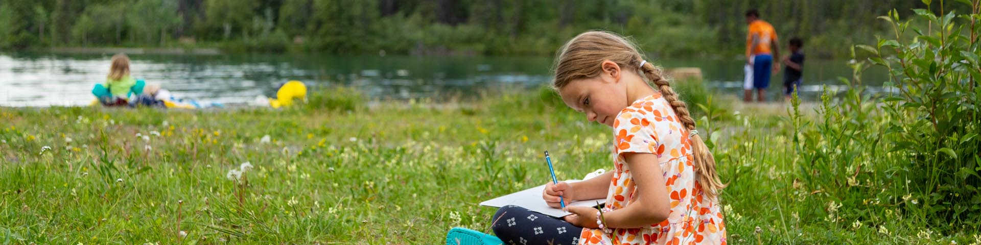 A young girl sitting in the grass writing in a notebook at Kathleen Lake with visitors and mountains in the background, at Kluane National Park and Reserve.