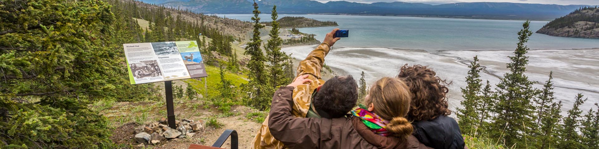 Three visitors on a bench snap a selfie in front of Kluane Lake on the Soldier's Summit Trail in Kluane National Park and Reserve.