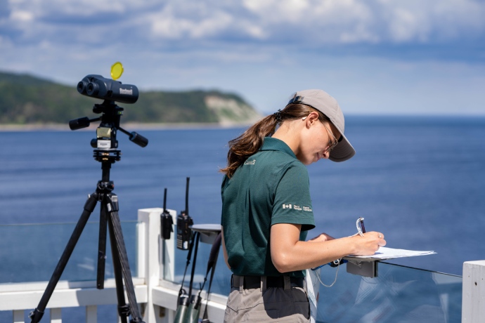 A Parks Canada staff person stands on a water viewing deck next to mounted binoculars while looking down to write on a clipboard.